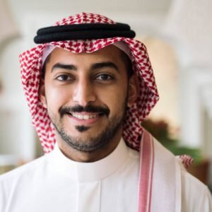 Head and shoulder view of Middle Eastern man wearing traditional dish dash, kaffiyeh, and agal and smiling at camera.