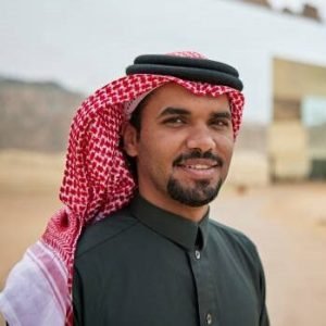 Headshot of bearded man in traditional clothing smiling at camera with global cultural center, sandstone outcrops, and desert area in background.